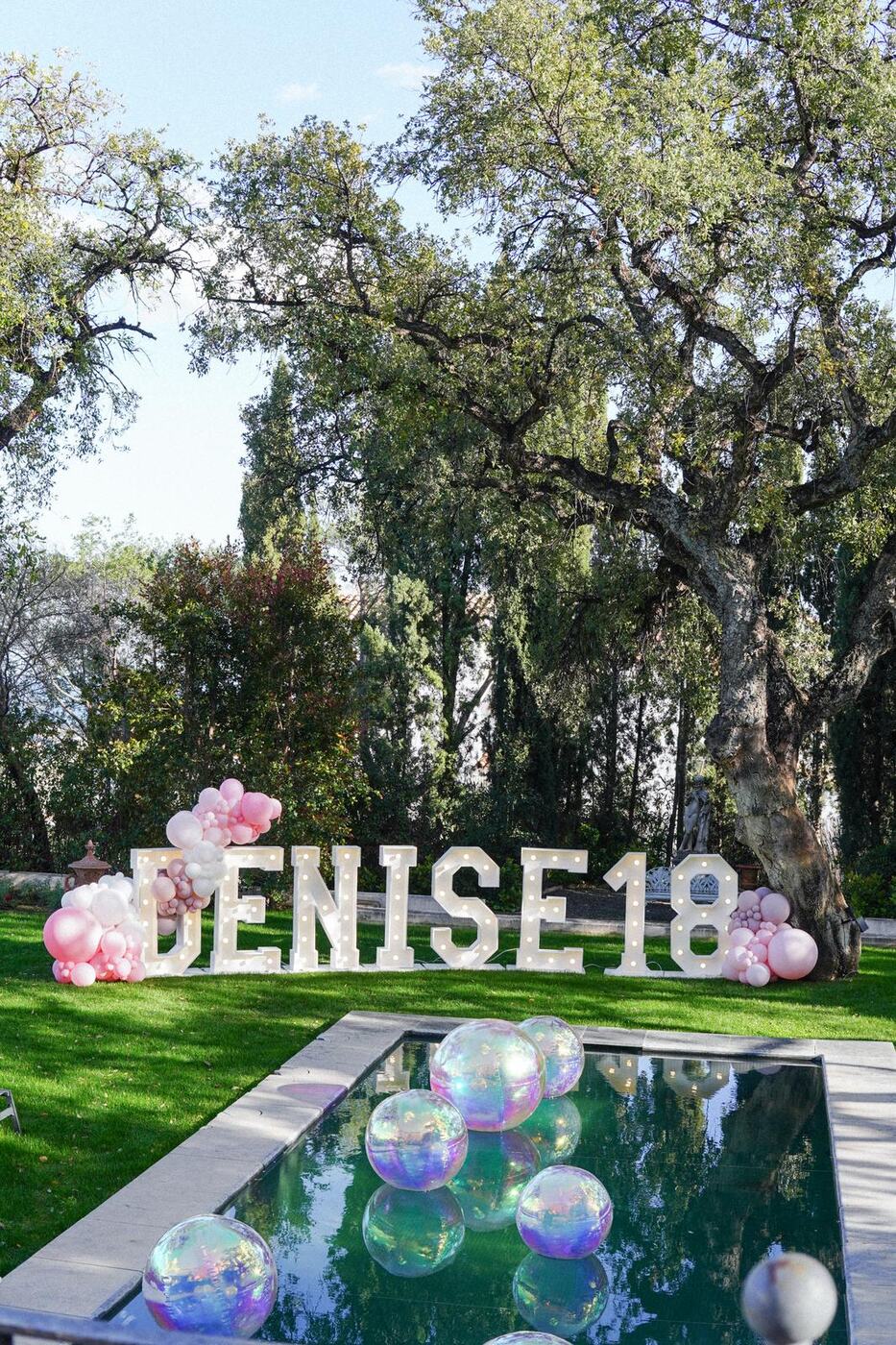 Light-up name letters and 18 numbers with pink balloon clusters and iridescent orbz floating in a pool, Marbella villa