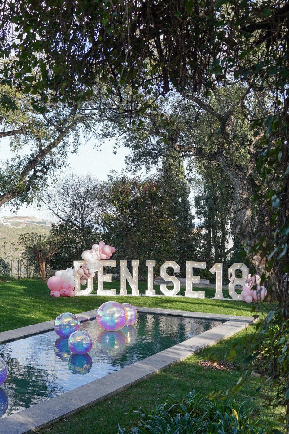 Iridescent orbz balloons floating in a pool beside light-up marquee letters, Marbella garden party